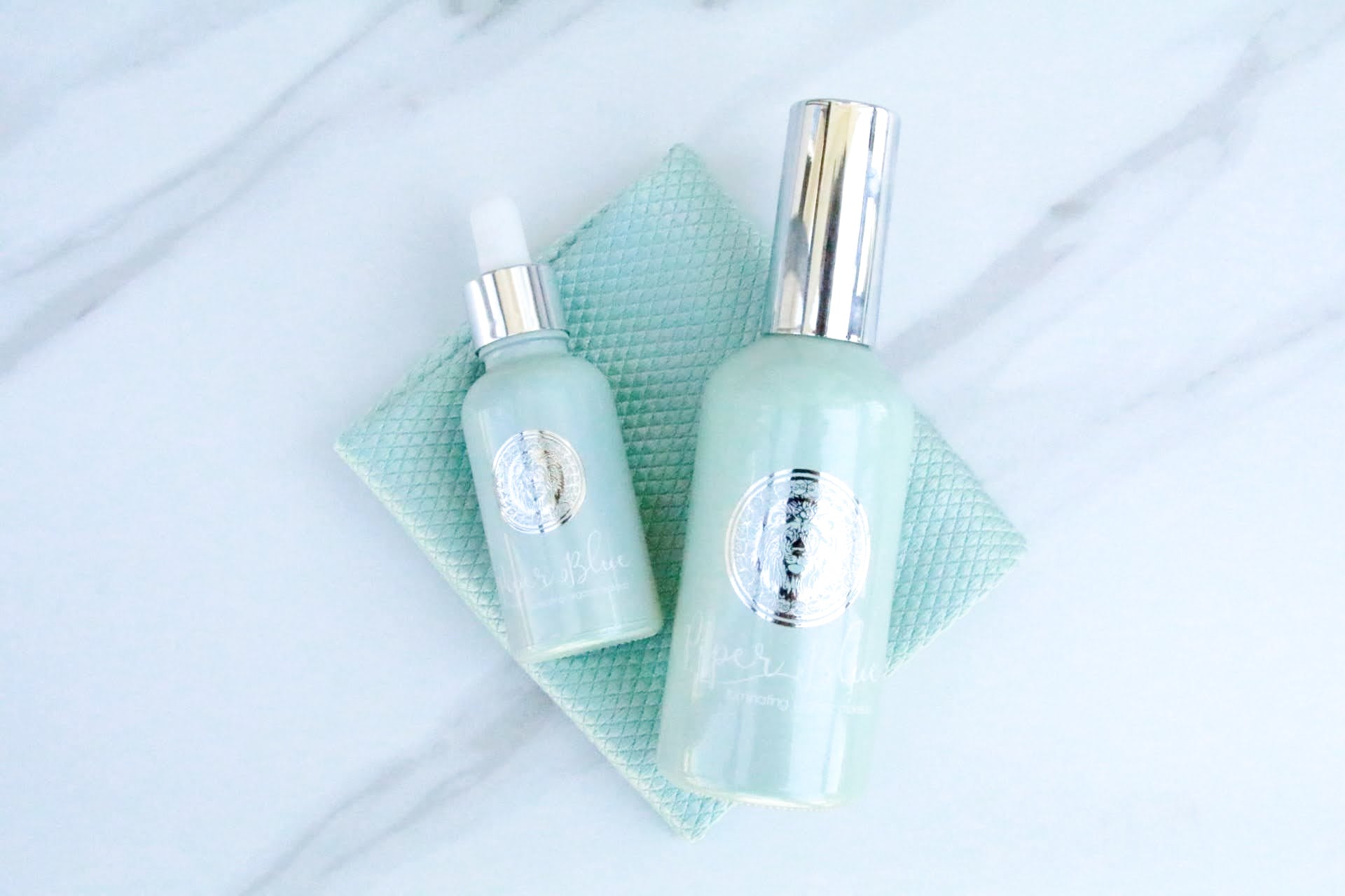 Blue glass bottles of Ageless Skin Care Flawless Beauty Serum and Setting Spray on an organic blue makeup removal cloth