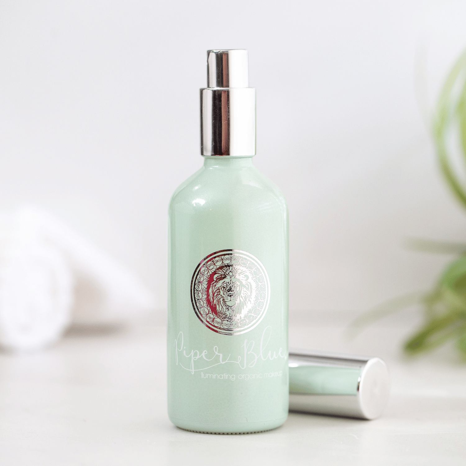 A bottle of Ageless Setting Spray with a green tint. The cap of the bottle is silver, and it is displayed on a light-colored surface with a plant in the background.