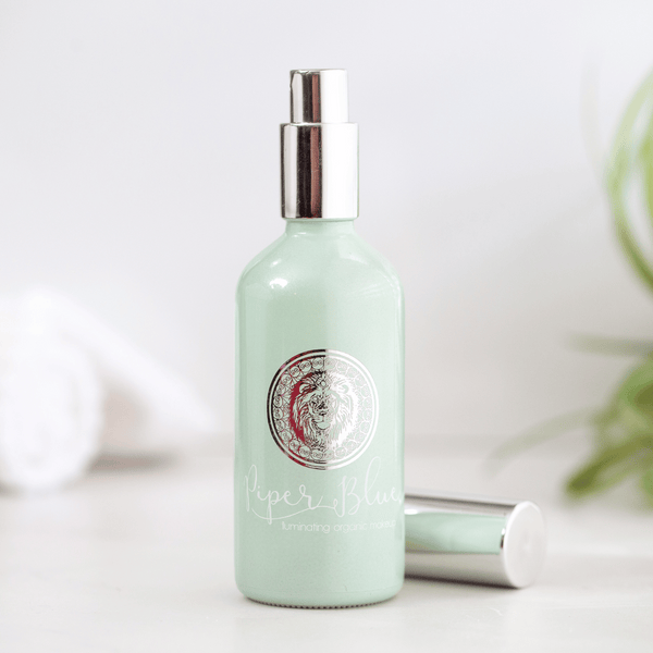 A bottle of Ageless Setting Spray with a green tint. The cap of the bottle is silver, and it is displayed on a light-colored surface with a plant in the background.