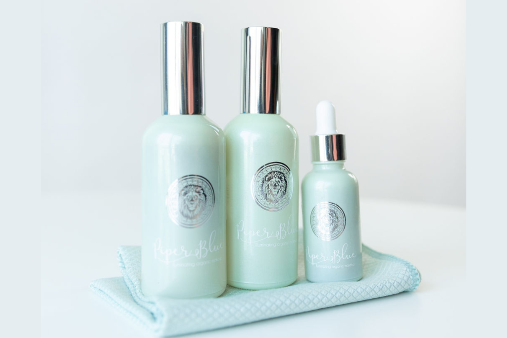 Two larger green toned bottles next to one smaller green toned dropper bottle set on a light blue makeup remover cloth with a white background.  Natural skin care set